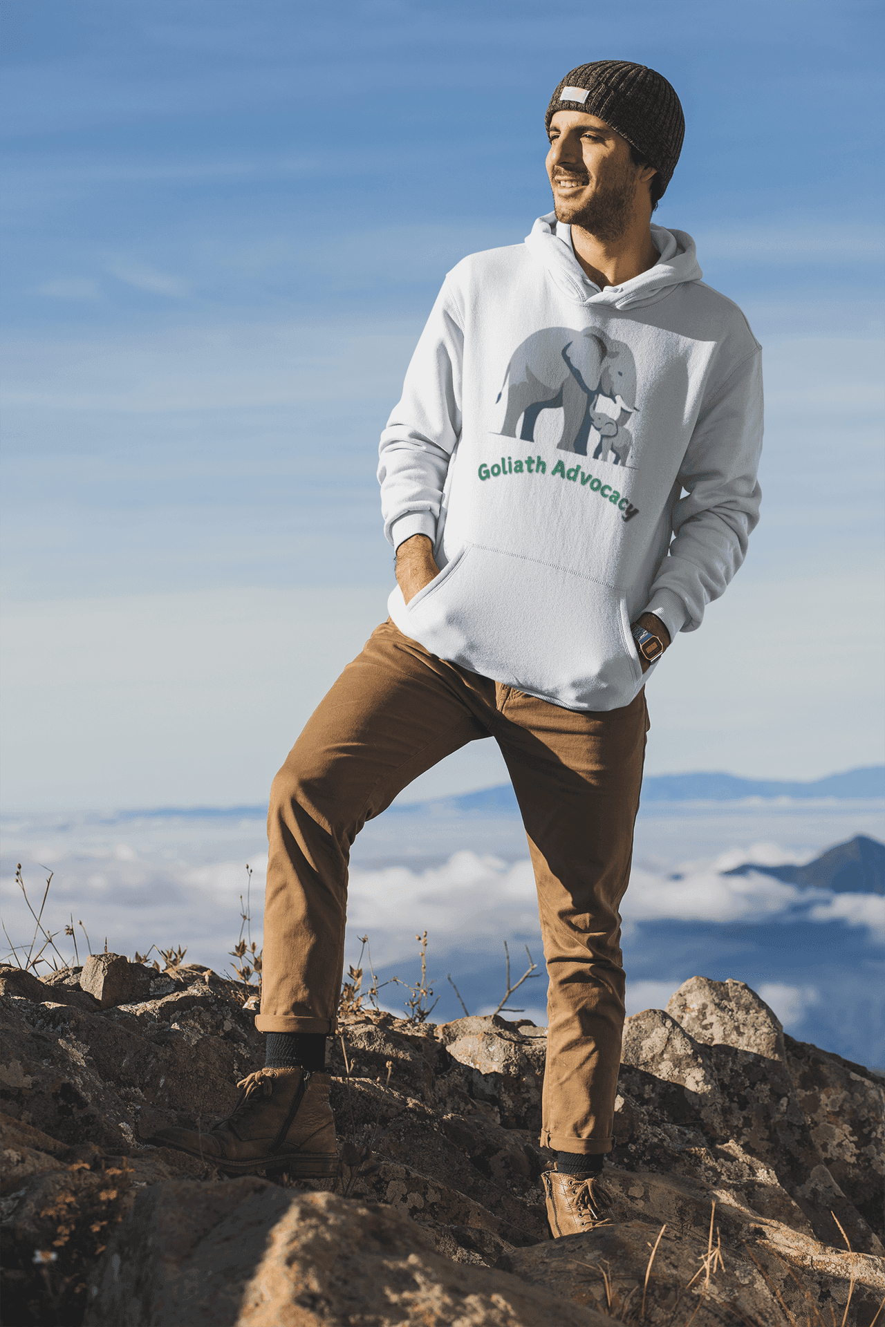 Person wearing Goliath Advocacy hoodie standing on rocky terrain against a backdrop of clouds and mountains.