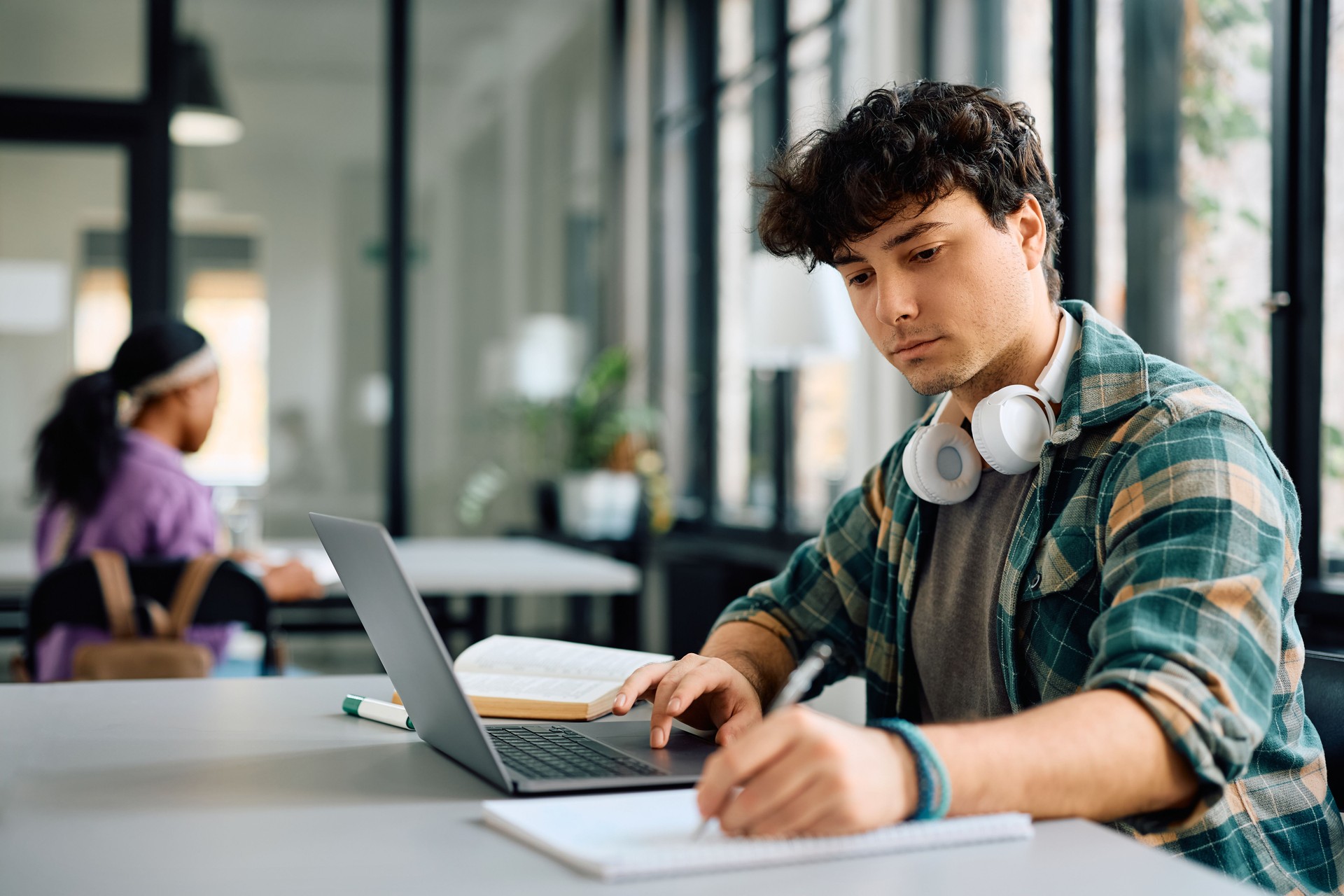 Young student taking notes while e-learning on laptop at the university.